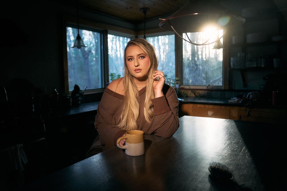 Young woman with long blonde hair sitting at a kitchen table holding a coffee mug, illuminated by soft natural light from large windows, creating a warm and inviting atmosphere.
